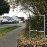 Foto des Dorfeingangs von Nackenheim kommend, zu sehen ist der leere Metallrahmen, in den das renovierte Eingangsschild wieder montiert wird.
