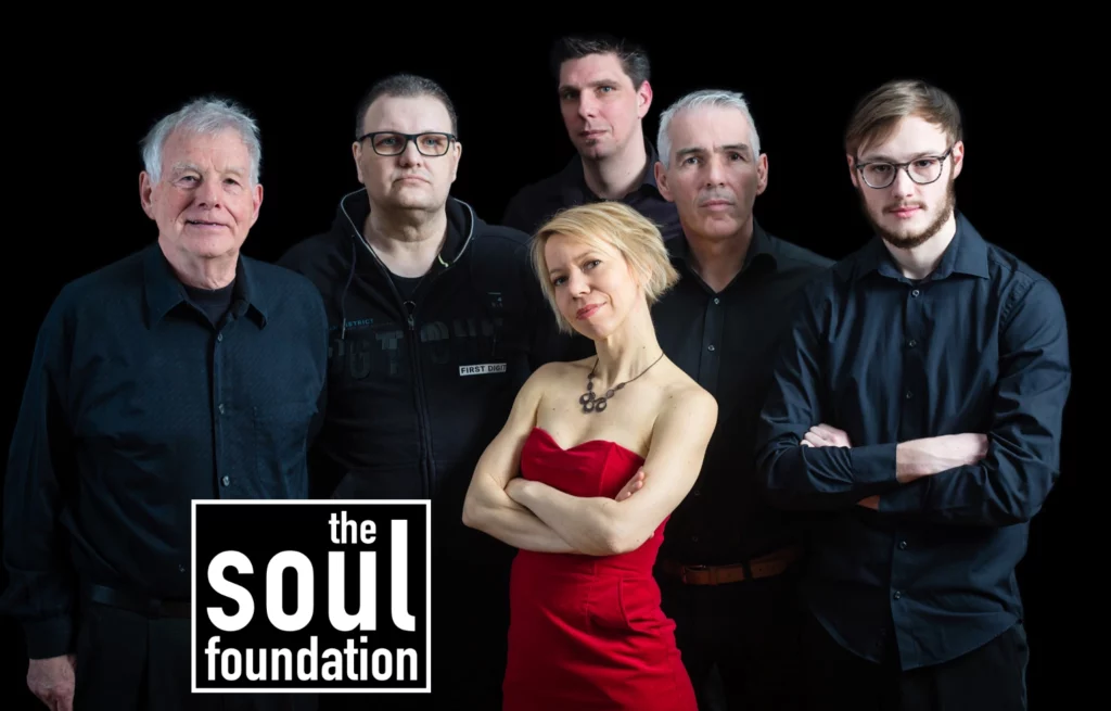 Die Band mit fünf Männern vor schwarzem Hintergrund und vor ihnen eine Frau im roten Trägerlosen Kleid. links unten der Schriftzug the soul foundation. 