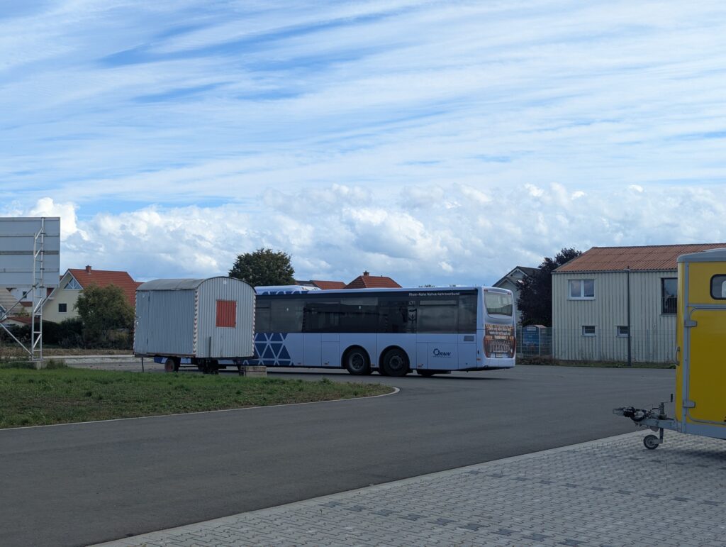 Ein Bus mit weiß-blauer Deko steht auf dem Parkplatz der neuen Halle, die Malteserhalle im Hintergrund.