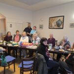 Ein weiß gestrichener Raum mit einem quer stehenden großen Tissch. Am Tisch sitzen 10 Frauen an einer Kaffee- und Kuchentafel. Hinter dem Tisch stehen ein größerer mittelalter Mann mit Brille und Bart und eine weitere Frau. Die Frau hat ein blaues Plakat in der Hand.