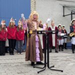 Ein Mann in einem Brau-rosa Kleid und dick geschminkt steht an einem Stehtisch und hat die Arme wie zur Segnung erhoben. dahinter das Jugendkomitee und das Komitee