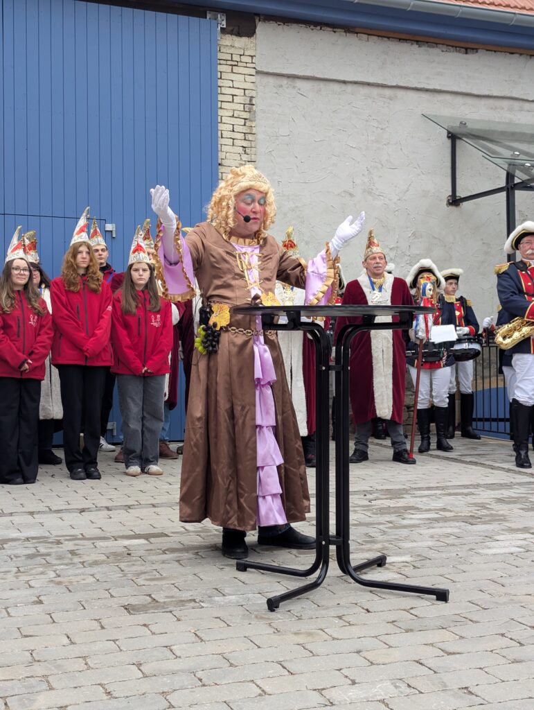 Ein Mann in einem Brau-rosa Kleid und dick geschminkt steht an einem Stehtisch und hat die Arme wie zur Segnung erhoben. dahinter das Jugendkomitee und das Komitee