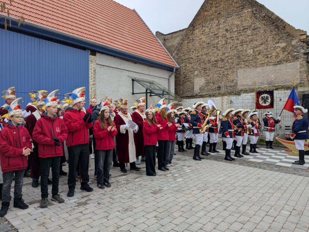 Vor der Scheune auf dem Dorfplatz steht links das Komitee und das Jugendkomitee, rechts daneben die Schoppengarde in ihren Uniformen und mit Instrumenten.