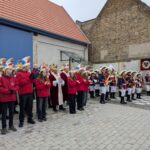 Vor der Scheune auf dem Dorfplatz steht links das Komitee und das Jugendkomitee, rechts daneben die Schoppengarde in ihren Uniformen und mit Instrumenten.