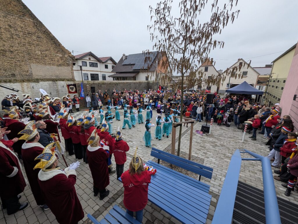 Blick von einer Treppe, links steht das Komitee, davor tanzen Mädchen in türkisenen Kostümen, davor schaut eine Menschenmenge zu.