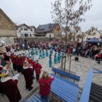 Blick von einer Treppe, links steht das Komitee, davor tanzen Mädchen in türkisenen Kostümen, davor schaut eine Menschenmenge zu.
