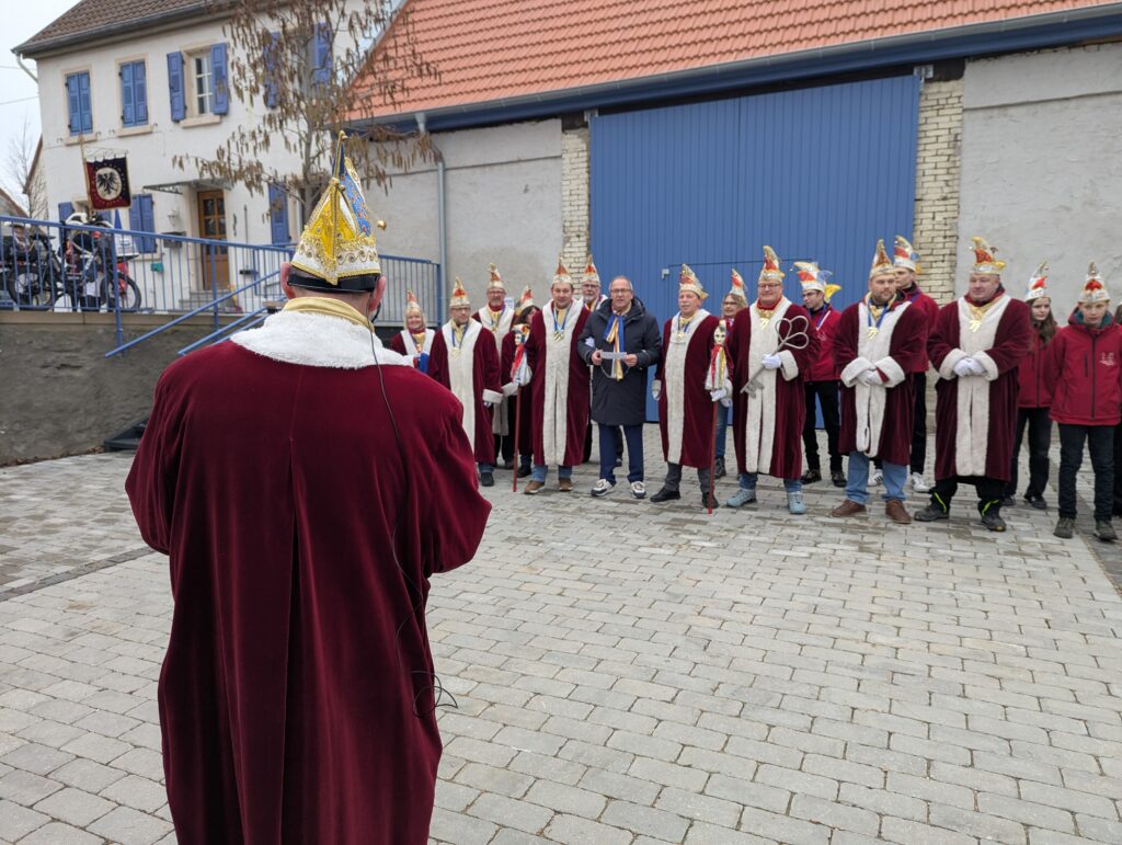 Links im Bild ein Mann von hinten in einem roten Mantel mit Pelzkragen und einer Narrenkappe, ihm zugewandt steht eine Gruppe von weiteren Männern mit den selben roten Mänteln und Kappen. Einer hält einen großen eisernen Schlüssel. dazwischen ein Mann in blauer Kleidung und mit einer Kette um die Hände.