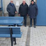 Drei Männer vor einer Wand und einem Schild mit der Aufschrift 'Babbelbank' (Rest nicht zu lesen). Vor ihnen eine Tisch-Bank-Garnitur in mittelblau