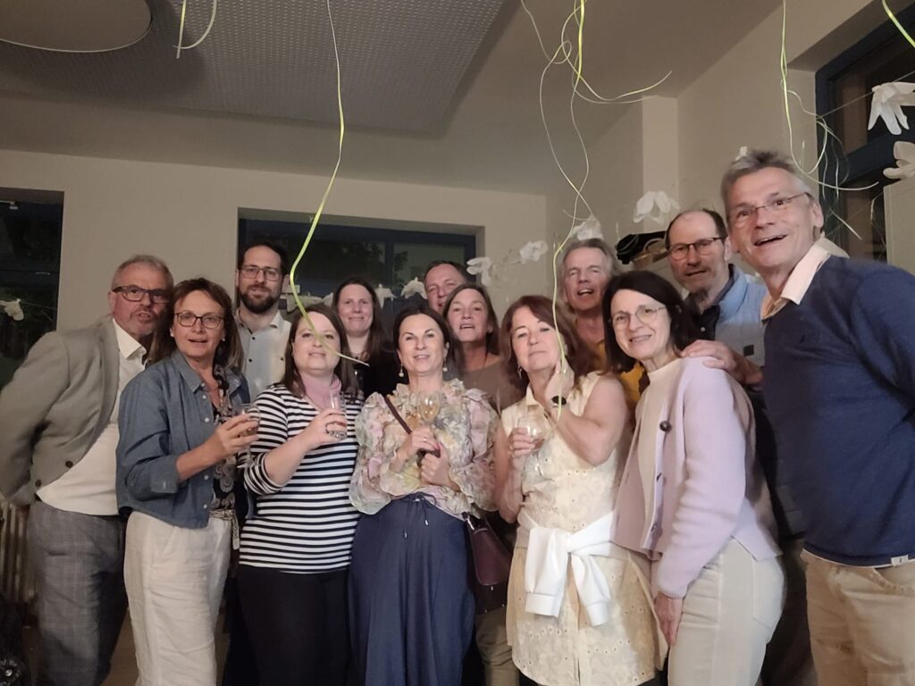 Eine Gruppe von Menschen steht in einem Raum, einige haben Weingläser in der Hand, von der Decke hängen Fäden von Luftballons