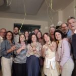 Eine Gruppe von Menschen steht in einem Raum, einige haben Weingläser in der Hand, von der Decke hängen Fäden von Luftballons
