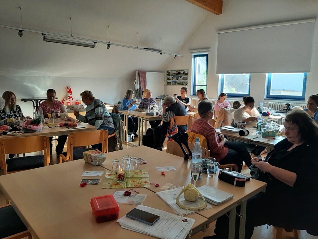14 Frauen sitzen an Tischen in einem hellen Raum und arbeiten an Handarbeitsprojekten. Einige haben Wolle und Stricknadeln in der Hand, manche unterhalten sich.