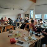 14 Frauen sitzen an Tischen in einem hellen Raum und arbeiten an Handarbeitsprojekten. Einige haben Wolle und Stricknadeln in der Hand, manche unterhalten sich.